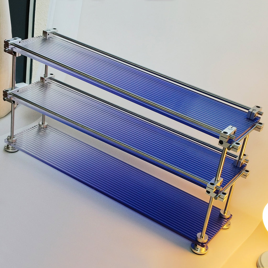 High-angle shot of HushLume 3-tier chrome steel linear shaft desktop storage shelf with fluted blue acrylic panels, showing industrial mechanical details.