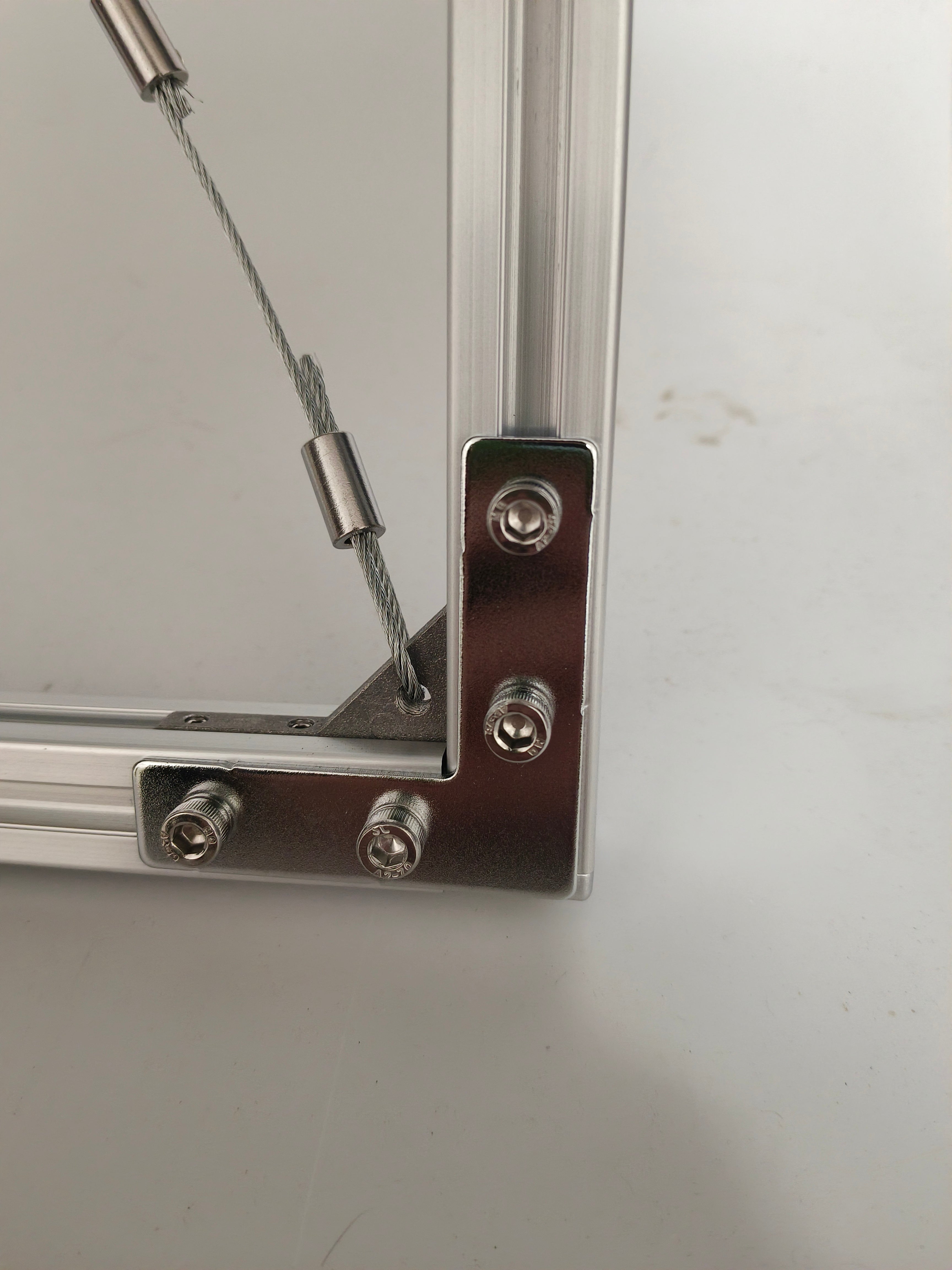Macro close-up of HushLume industrial aluminum floating frame corner joint, detailing the stainless steel wire rope, crimp sleeve, and heavy-duty hex screws.