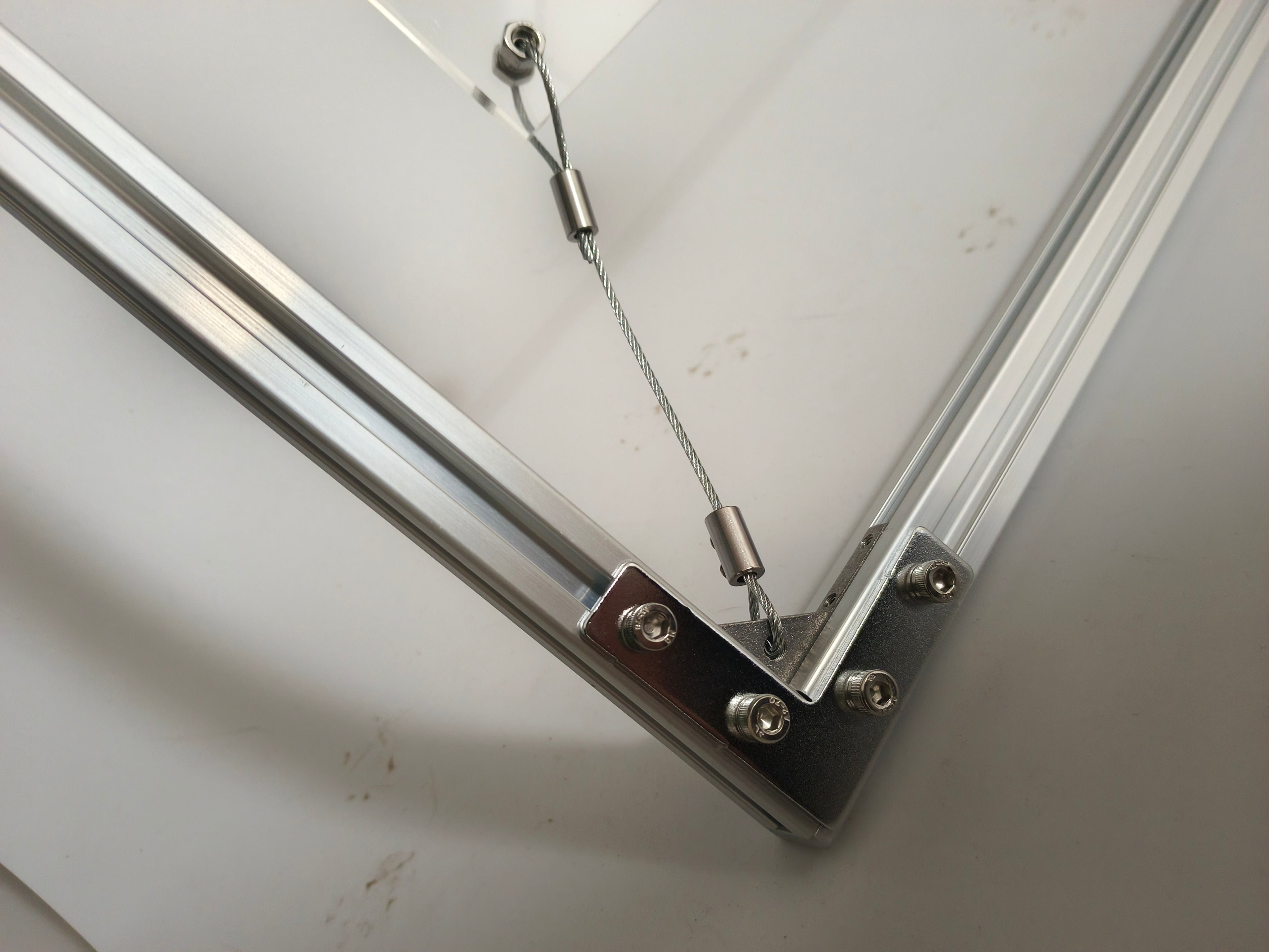 Angled close-up of HushLume industrial floating frame, showing the stainless steel tension wire fixing method on the clear acrylic panel corner.