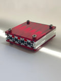 Close-up product shot of HushLume 10x10cm industrial aluminum coaster featuring vibrant translucent ruby red acrylic plates in natural sunlight.